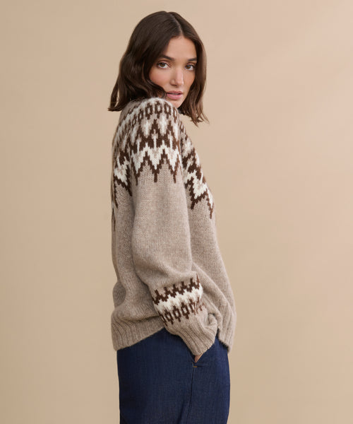 A woman with shoulder-length brown hair wears the Thatcher Crewneck, a beige alpaca blend sweater with a dark brown and white fair isle yoke, paired with blue jeans. She stands against a beige background, looking over her shoulder at the camera.