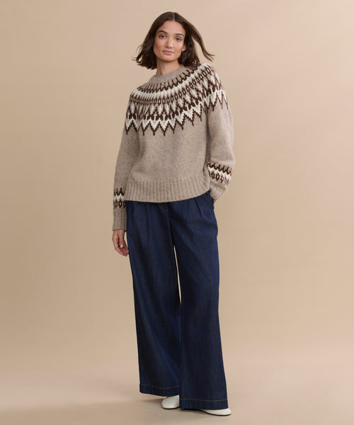 A woman with shoulder-length brown hair and a neutral expression stands against a beige background wearing the Thatcher Crewneck sweater, wide-leg dark blue jeans, and white shoes.