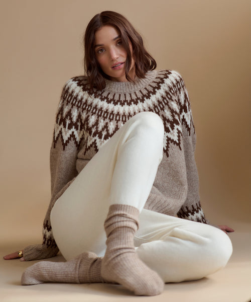 A woman with wavy brown hair sits on the floor against a beige background, wearing the Thatcher Crewneck sweater, white pants, and beige socks. She looks at the camera with a relaxed expression.