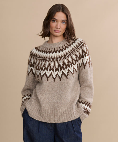 A woman with shoulder-length brown hair wears a beige Thatcher Crewneck sweater and dark blue pants, standing against a plain beige background.